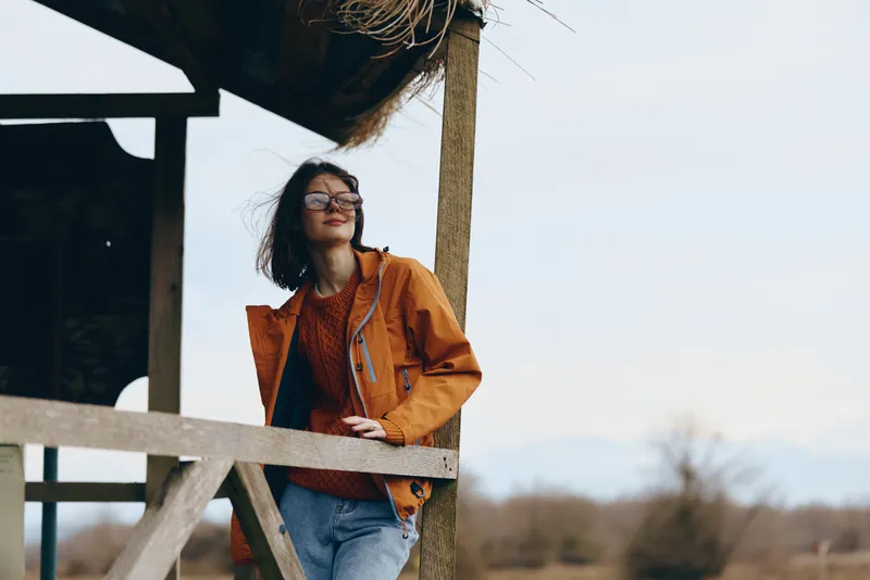 Woman lifestyle outdoors in spring, orange jacket and smiling by wooden fence, glasses on, relaxed pose in breeze, sunlit daylight, blurred countryside field background