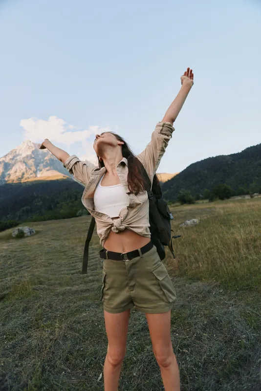 Young woman stands in a grassy meadow with a mountain backdrop, arms raised in triumph, creating a bright, energetic outdoor moment.