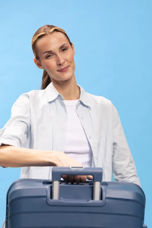 Young woman with casual outfit holding blue suitcase on light blue background. Travel concept, vacation preparation, and summer trip planning with confident and relaxed mood.