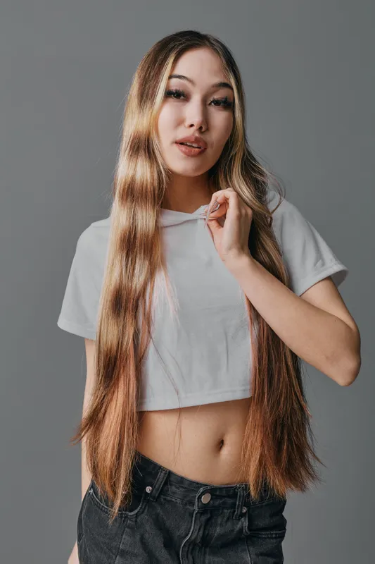 Young woman with long straight hair wearing cropped white t shirt and black jeans posing on gray background with confident expression and natural makeup.