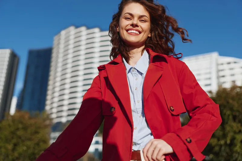 Young woman in a red coat smiles broadly as she stands outdoors with modern city buildings behind, exuding confident, lively street style and a sunny, upbeat mood on a clear day.