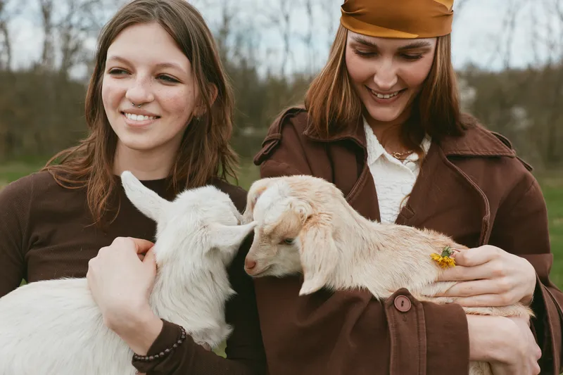 Cheerful women outdoors, two friends cuddle friendly goats, sunlit countryside moment, relaxed posture, natural colors, gentle breeze, candid interaction, rustic clothing, lighthearted lifestyle scene