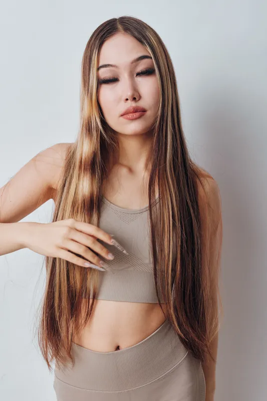 Young woman with long straight hair and closed eyes wearing a sleeveless top and matching pants posing in front of plain light background indoors in natural light.