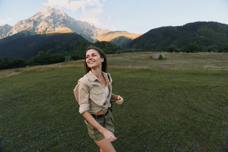 Young woman exploring a grassy field with towering mountains in the background, smiling as warm sun highlights her casual outfit and carefree mood