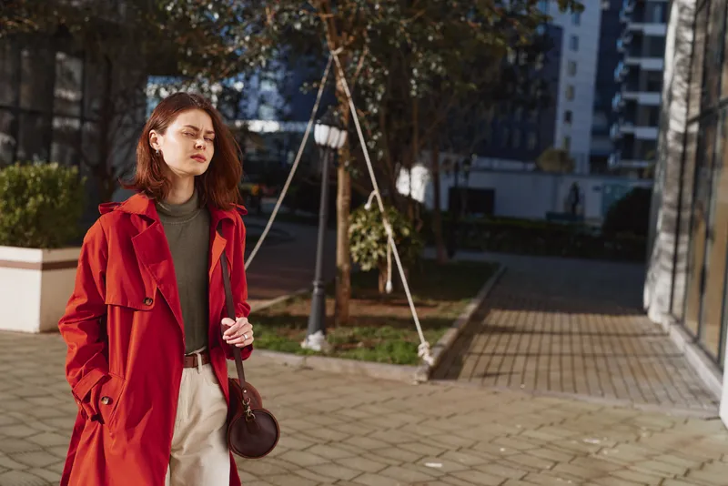 Young woman with red hair wears a red trench coat and beige pants while walking confidently on a sunny urban street with modern buildings in the background.