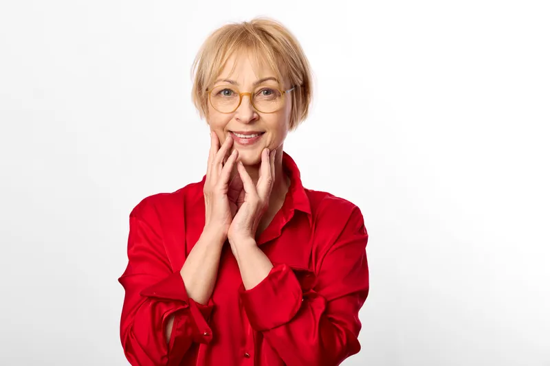 Woman senior in red shirt smiling, hands near face, closeup studio portrait on white background, friendly gesture for beauty grooming, skincare wellness and calm confidence, soft light