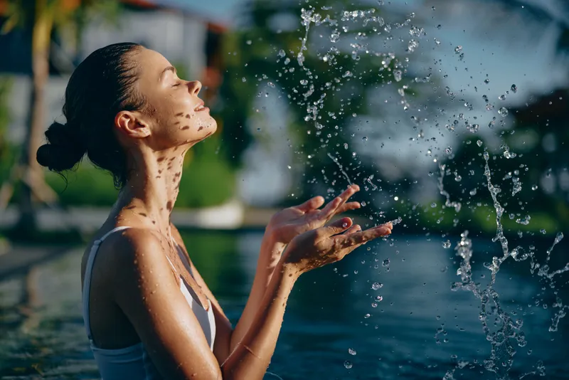 A woman enjoys a refreshing moment by a pool, water spraying gently as sunlight warms her face, outdoor air bright and crisp. A lifestyle scene focusing on leisure, wellness, and joyful release.