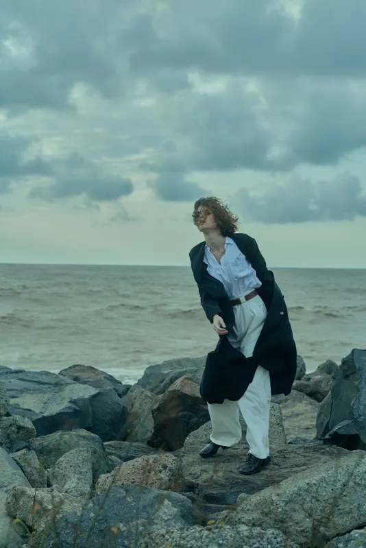 Individual walking along a rocky shoreline in a long coat and white shirt, wind-swept hair, moody ocean backdrop, cloudy sky, dynamic movement, fashion inspired scene of solitude and resilience