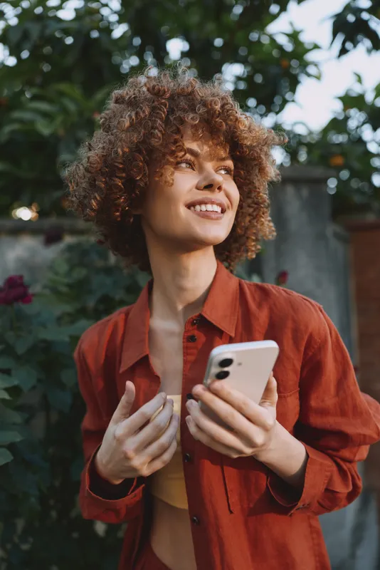 Young woman with curly hair smiles while holding a phone outdoors, dressed in a casual rust shirt, enjoying a relaxed moment in a sunlit garden