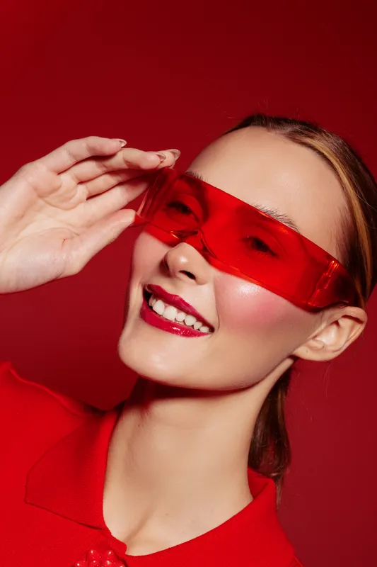 Smiling woman with red transparent glasses and red lipstick poses on red background. Fashion portrait with vibrant colors and confident expression. Concept of style and beauty.