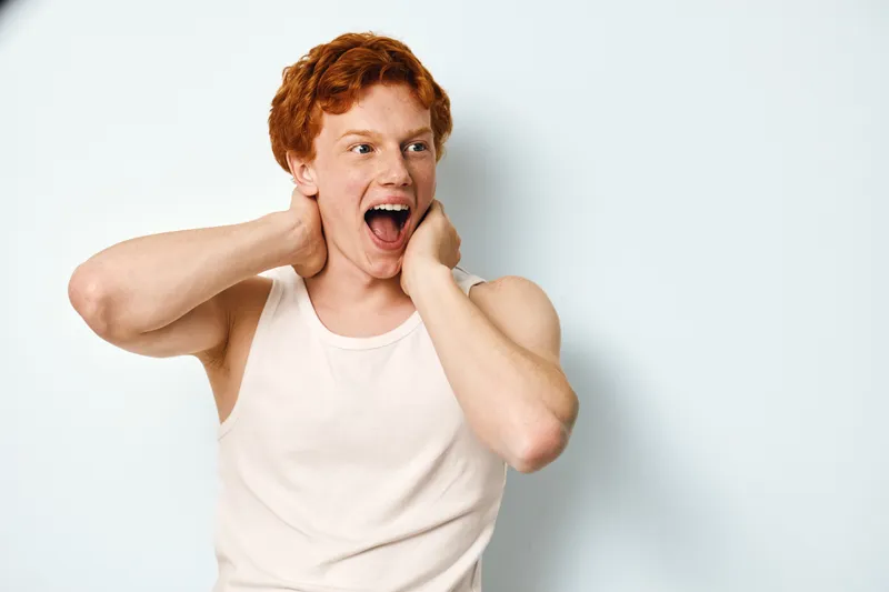 Young man with red hair in white tank top surprised expression with hands on neck looking sideways in studio with white background.