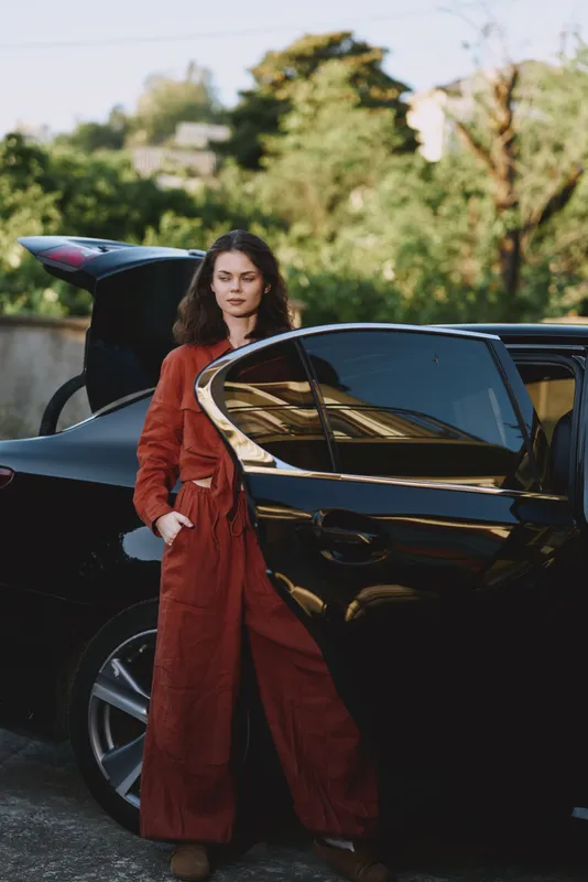 A stylish woman in a red jumpsuit stands beside a sleek black car, outdoors in daylight. A candid lifestyle moment emphasizing fashion, confidence, and urban wanderlust