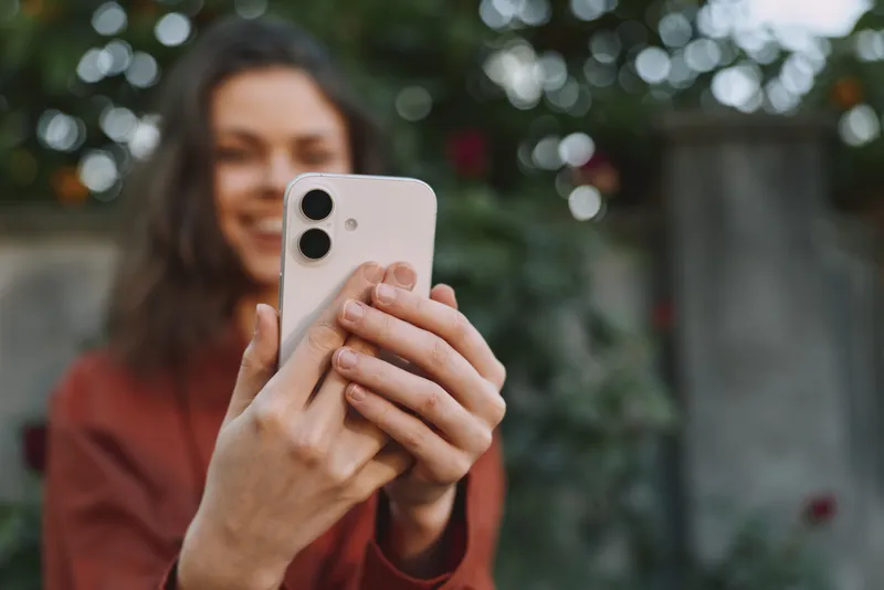 Smiling woman outdoors taking a selfie with a smartphone, casual lifestyle moment, warm natural light, lush greenery background, cheerful mood