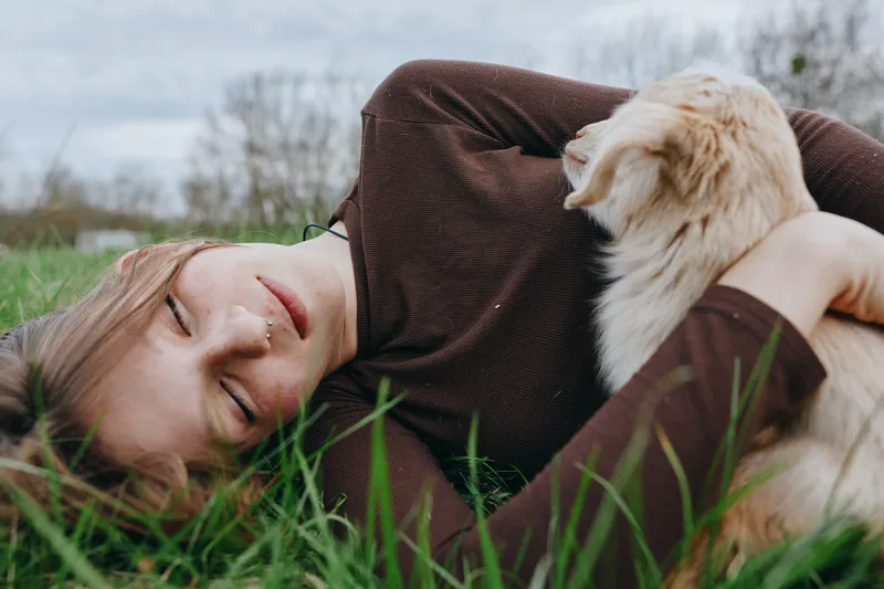 A relaxed woman lying on a grassy field outdoors, cuddling a small dog. A lifestyle moment highlighting comfort, calm, and companionship in a natural, sunny setting.