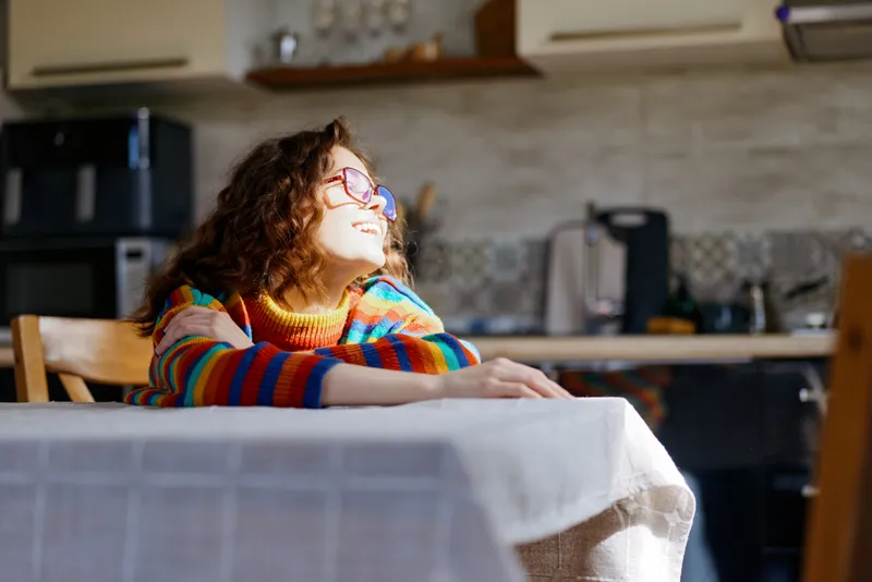 Cheerful woman indoors, sitting at a table in a sunlit kitchen, colorful striped sweater, relaxed pose, warm natural light, candid moment, home atmosphere, soft breeze, vibrant colors, everyday