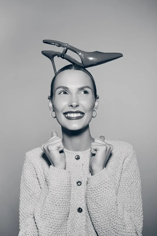 Smiling caucasian woman with light skin and earrings holds collar of cardigan with both hands and balances shoe on head, studio portrait.