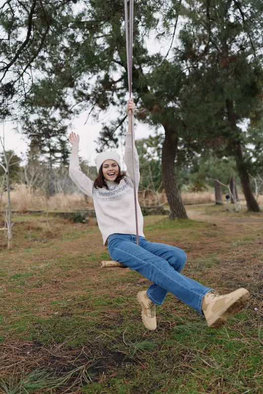 Girl on a swing in a sunny outdoor park, wearing a casual sweater and jeans, smiling as she sways among trees and grass in a carefree vibrant moment
