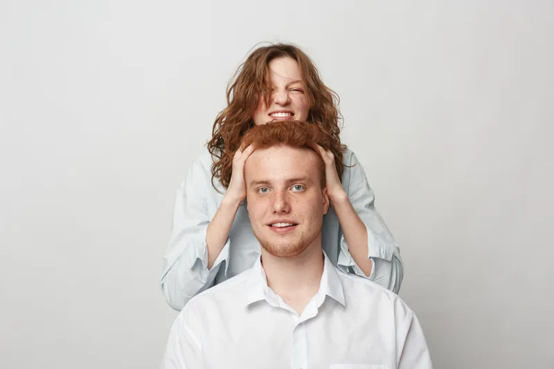 Smiling young woman with curly hair playfully covers the eyes of a redhead man in white shirt. Studio portrait on white background. Concept of friendship, fun, and surprise moments.