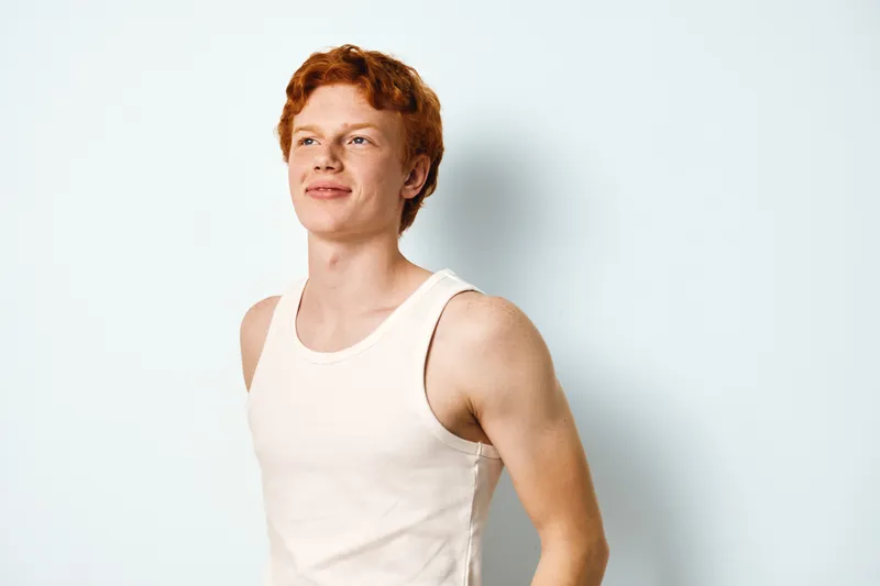 Young man with red hair wearing white tank top looks confident and relaxed standing against light background in casual style and natural pose indoors.