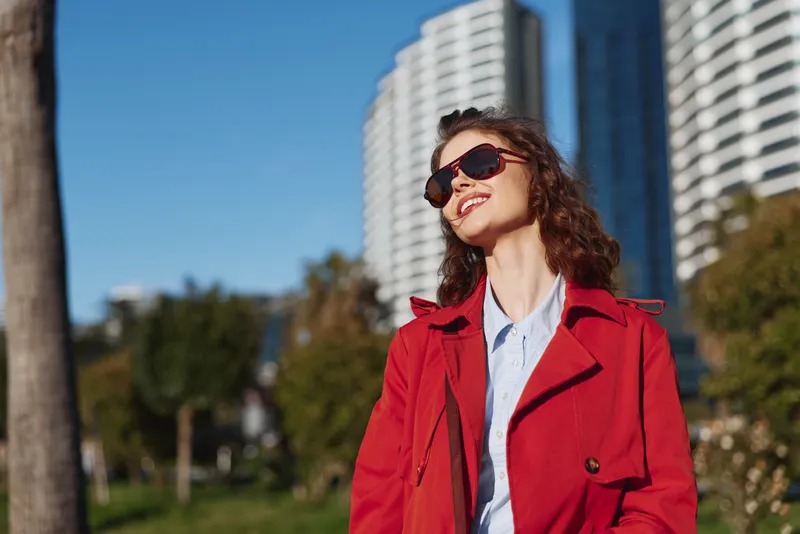 Young woman with curly hair wearing a red coat and sunglasses smiles while enjoying a sunny day outdoors in an urban park setting.