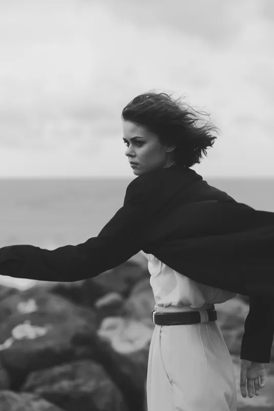 Female model with cape and windswept hair on a rocky coast. Monochrome seascape setting, moody and dramatic fashion mood, minimalist composition, ocean horizon, graceful pose, timeless elegance in