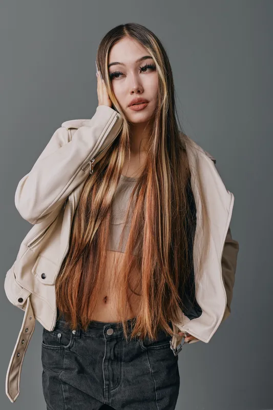 Young woman with long hair wearing stylish jacket and casual top posing against gray background. Fashion portrait of confident female model in modern outfit and natural makeup.