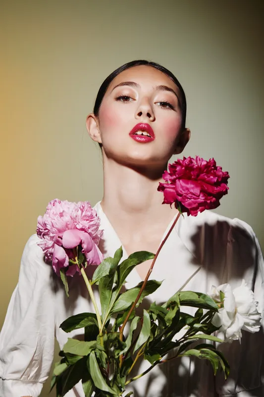 Young woman with flawless makeup poses with bright pink peonies against beige backdrop. Soft shadows create depth. Concept for beauty creativity and fashion editorial shots.