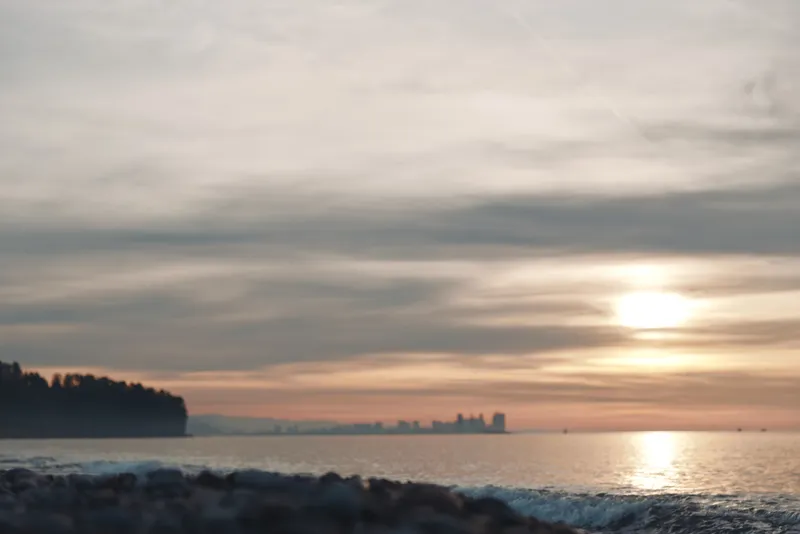 Sunset over a calm ocean with distant skyline, soft clouds and warm reflections on water, tranquil beach foreground and gentle waves at dusk.