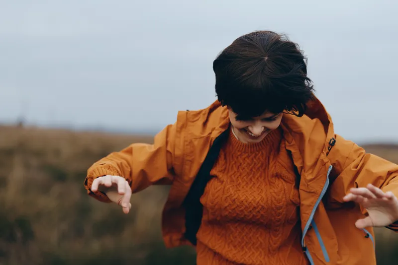 Woman lifestyle outdoor autumn, orange sweater and jacket, smiling while walking in a grassy field, hands adjusting sleeves, hair blowing, warm daylight, nature landscape, countryside sky