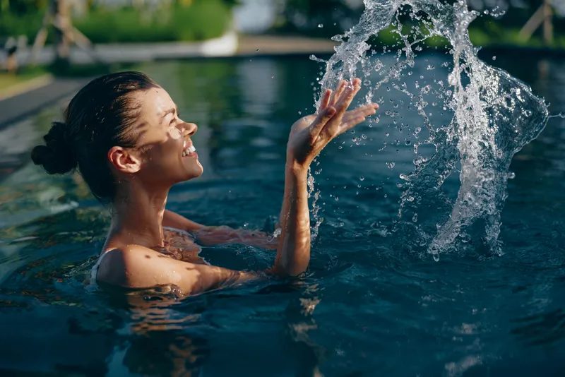 Woman in pool enjoying a bright splash, smiling as water arcs from her hands, outdoors, relaxed lifestyle moment of play and wellness in a calm blue pool setting