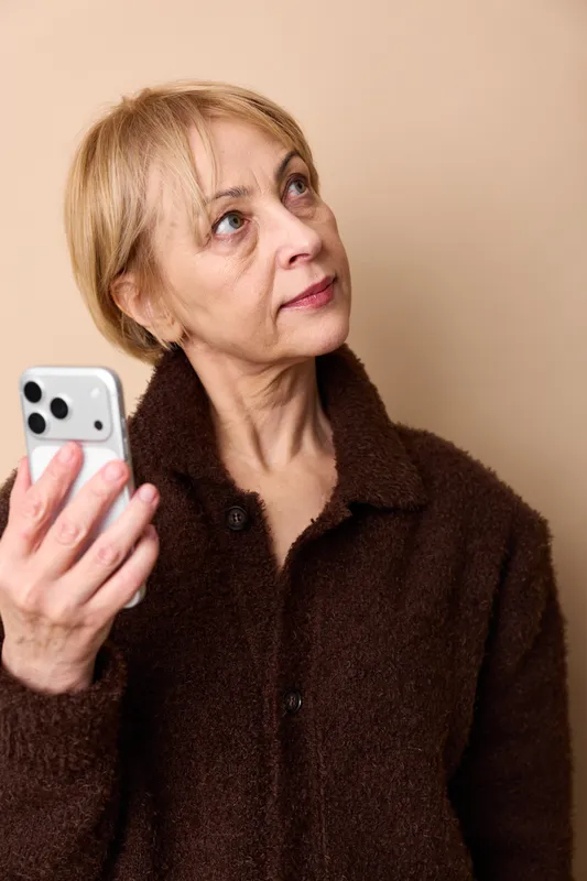 Woman senior smartphone portrait indoor communication texting browsing technology casual jacket, elderly woman holding phone, thoughtful face, warm indoor light, closeup, relaxed posture