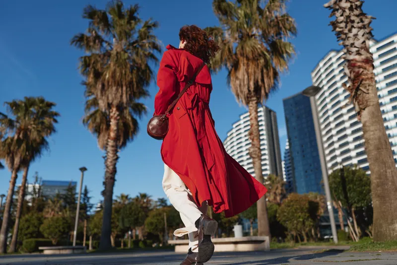 A woman in a bright red trench coat strides along a palm lined urban boulevard under a clear blue sky, showcasing bold street style and energetic movement as passersby observe.