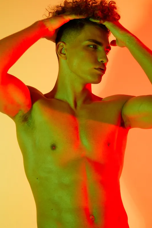 Young muscular man posing shirtless with hands on head under colorful red and green lighting, showing strong physique and confident expression in studio portrait.