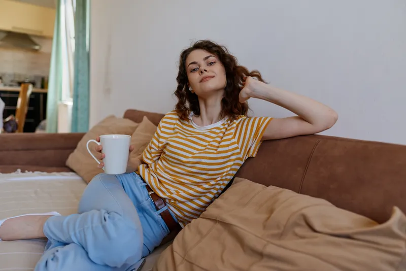 Cheerful woman relaxing at home on a couch, striped t shirt, casual jeans, coffee mug nearby, natural light, cozy living room, candid lifestyle moment