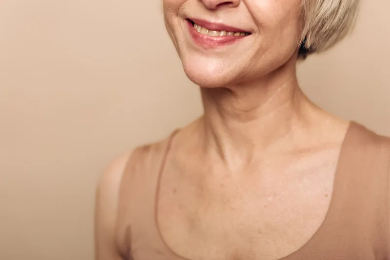 Closeup of mature woman smiling gently in beige tank top against neutral background. Natural skin texture and subtle makeup highlight graceful aging and calm positive mood.
