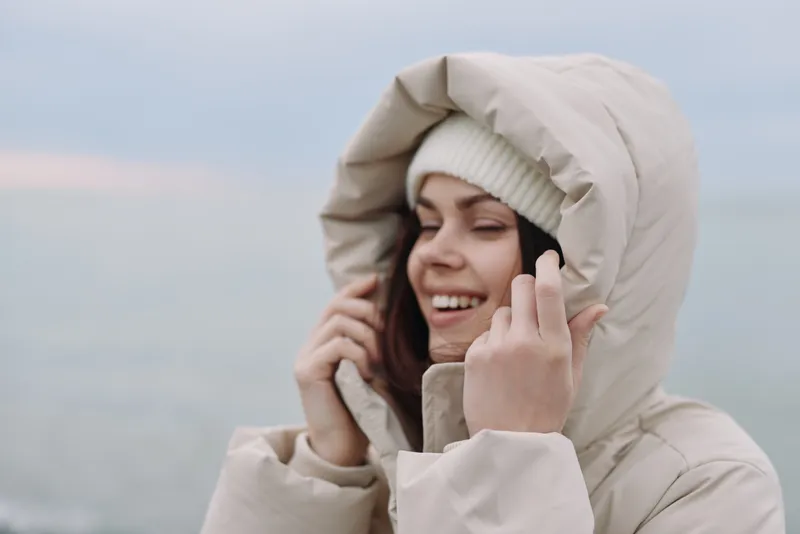 Winter, woman, outdoors, hooded jacket, smiling as she adjusts her beanie, cold breeze and soft ocean backdrop, gentle light, serene mood, cozy style for chilly days
