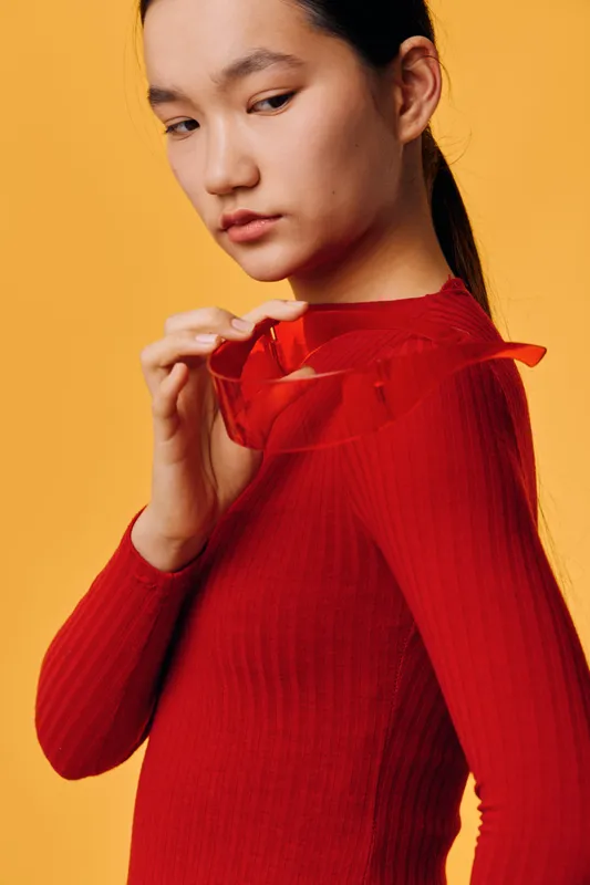 Woman in red sweater holding red sunglasses looking down with serious expression on orange background. Portrait, fashion style, casual wear, accessory, calm mood, model posing.