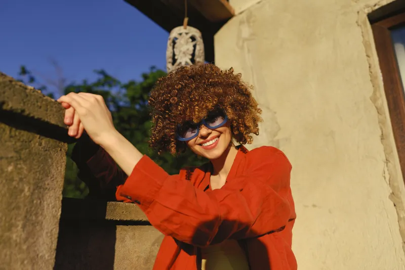 Smiling woman with curly hair leaning on a stone wall wearing a red jacket. Sunny outdoor scene emphasizing lifestyle, fashion, and a carefree mood