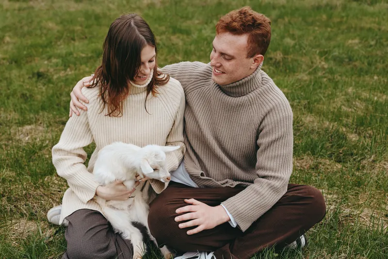 Smiling couple on the grass with a fluffy goat kid, cozy sweaters, soft natural light, lifestyle moment, family bond, gentle care, rural calm, connection and warmth