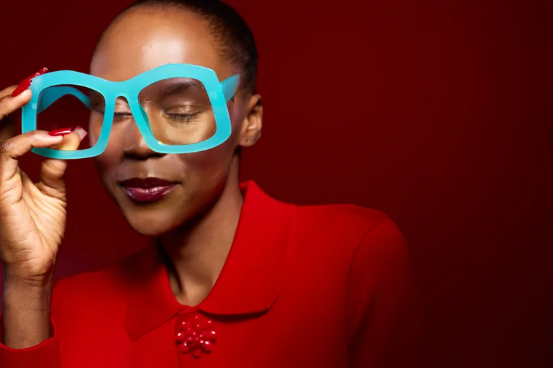Portrait of a confident black woman with dark skin wearing bright red clothing and holding oversized blue glasses on a deep red background. Concept for fashion and style.