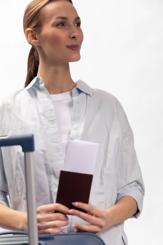 Young woman with passport and boarding pass stands next to suitcase, looking away. Travel concept for journey, vacation, planning, and airport security with casual clothing and natural light.