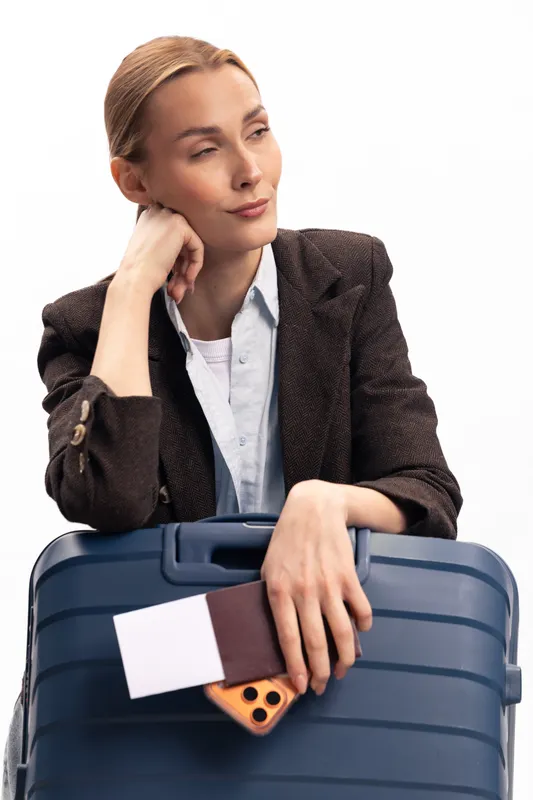 Thoughtful woman in blazer holding suitcase and passport with smartphone. Travel concept, waiting for flight or trip, business or leisure journey, isolated on white background.