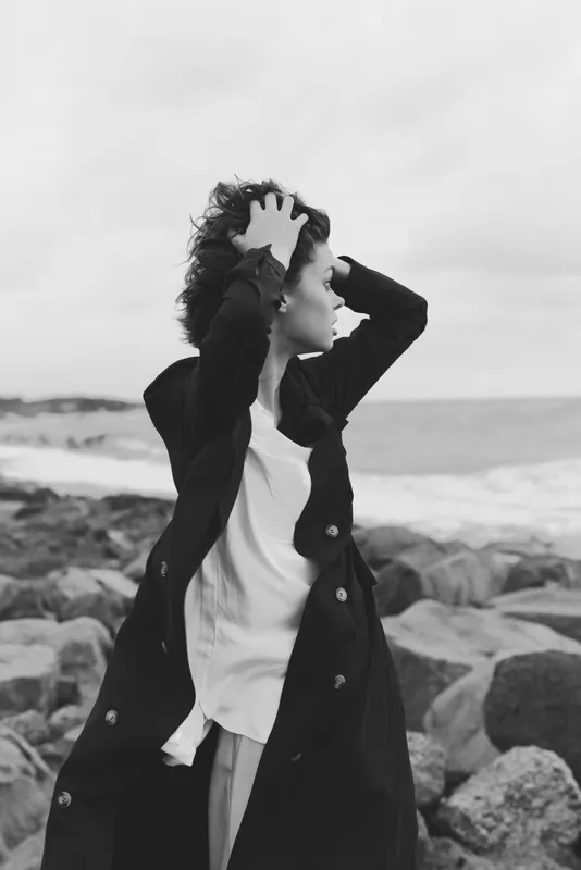 Beach wind scene with a windblown coat and contemplative woman on a rocky shore, monochrome mood, fashion and movement by the sea for artistic impact