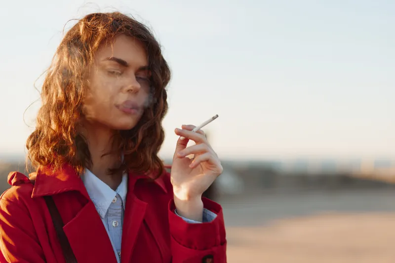 Young woman with curly hair smoking cigarette outdoors during sunset, wearing red coat and white shirt, soft natural light and blurred background.