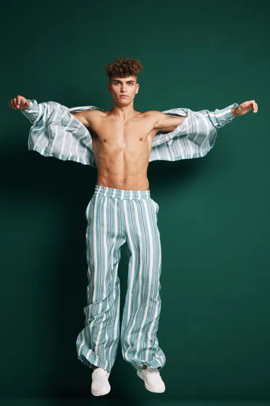 Young man with curly hair wearing striped pants and open shirt posing with arms outstretched against green background. Fashion, casual style and confident expression in studio.