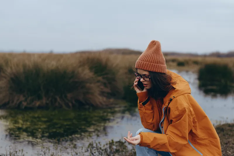 Woman in winter outdoors, wearing knitted hat and yellow coat, stands by river and smiles softly, lifestyle portrait in natural landscape with cloudy sky