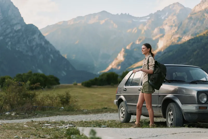 Young woman with a backpack stands next to a car on a mountain road enjoying the scenic view during a hiking trip in warm weather.