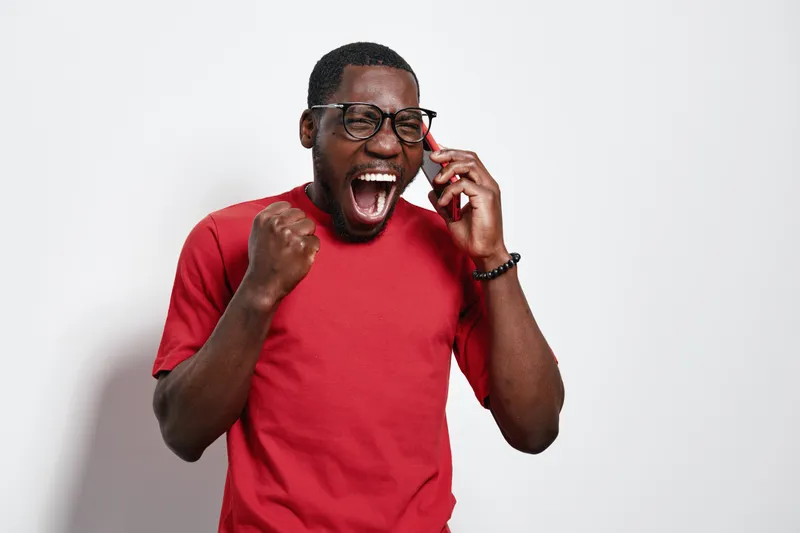 African American man with dark skin wearing glasses and red shirt talking on phone with excitement and clenched fist for success and positive news on white background.