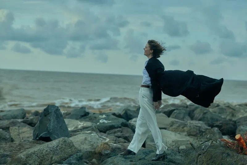 Businesswoman walks along a rocky shoreline with a flowing cape, blazer and trousers, amid a moody sea breeze and clouds, expressing determination, resilience, and coastal ambition