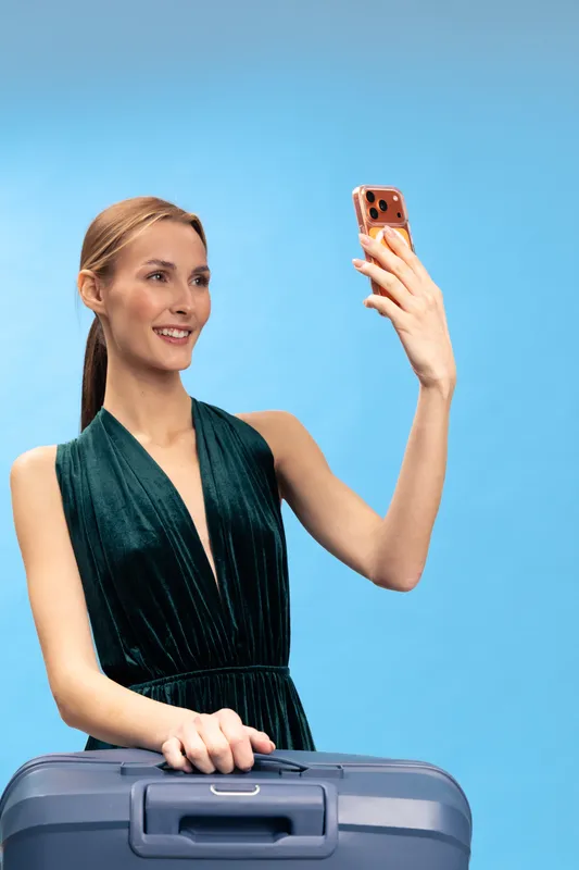 Young woman in elegant dress smiles while taking a selfie with smartphone. She holds a blue suitcase handle, posing against a plain blue background. Concept of travel and social media.